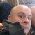 Pixelated close-up of a person with light skin, bald head, and dark clothing, facing slightly right with a neutral expression.