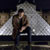 Pixelated static image of a person in dark clothing seated on a chair with a large light-colored structure behind them.