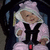 A child in a pink hooded outfit sits secured by black straps with red buckles, smiling slightly while seated in a dark blue car seat.