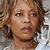 Pixelated close-up of a person with curly brown hair, light skin tone, and dark eyes, wearing a white top with a dark collar, against a blurred background.