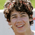 Pixelated close-up of a person with dark curly hair smiling, showing teeth, against a blurred background with green and light blue tones.