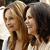 Two women with long hair stand side-by-side, one with light brown hair and the other with dark brown hair, both wearing sleeveless tops.