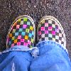 Static close-up of colorful checkered slip-on shoes on a person's feet wearing blue denim jeans, viewed from above against a dark textured background.