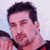 Pixelated static image of a person's face with dark hair and light skin, wearing a black and red garment against a pale blue background.
