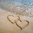 A beach scene with two heart shapes drawn in sand, a person's arm with a pink heart tattoo, and bold text reading "IM IN LOVE" on white background.