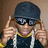 Pixelated image of a person wearing dark sunglasses, a black cap, and multiple gold chains while adjusting the cap with both hands.