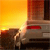 A static image shows a car at sunset with a bright yellow-orange sky and a red taillight visible on the vehicle.
