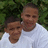Two people in white shirts stand close together against a green leafy background, both smiling with eyes closed.