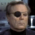 Static pixelated image of a person with short brown hair wearing a black eye patch over the left eye and dark clothing against a blurred background.