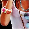 A close-up of a person's arm wearing a white bracelet with pink and green floral patterns, alongside a brown beaded necklace and a blue beaded necklace.