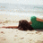 A person lies on a sandy beach with dark hair, wearing green clothing and brown shoes, facing the ocean under a light sky.