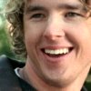 A close-up of a person with curly light brown hair wearing a dark shirt with a white collar, smiling broadly with visible teeth against a blurred green background.