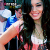 A woman with long dark wavy hair wears a pink and white short-sleeve top with layered necklaces, standing in front of a blurred crowd.