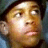 A pixelated close-up of a person's face with light skin tone, dark hair, and a neutral expression, set against a blurred background with blue and brown tones.