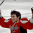 A hockey player in a red jersey raises arms with a stick while another player in white helmet and jersey looks left to right.