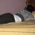 Pixelated static image shows a figure lying on a light brown surface with a muted purple background, wearing dark pants and a gray-and-white checkered shirt.