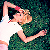 A person with blonde hair lies on green grass, wearing a white short-sleeve shirt and red wristband, with one arm raised above the head.