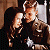 Two figures in close physical contact, one with light brown hair and the other with dark hair, positioned against a blurred background with warm tones.