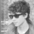 Black-and-white pixelated image of a person with dark hair and sunglasses, wearing a light-colored shirt, viewed from the side against a textured background.