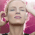 Pixelated image of a person with closed eyes, light skin, and blonde hair, wearing a pink top with dark straps, holding a pink object near their face.