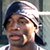 Pixelated static image of a person wearing a dark cap and light-colored headband with visible facial features including eyes, nose, mouth, and ear, set against a blurred background.