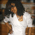 Woman with dark hair in white dress holding brown bag, static pose with slight head tilt.