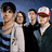 Four people stand together against a blue background; the right side displays red text reading "Dance Dance" in pixelated font.
