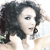 Pixelated close-up of a person with dark curly hair, light skin, and visible eye makeup, wearing a white top against a light background.