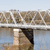 A static pixelated image shows a bridge with multiple arches spanning a body of water, reflecting in the calm surface below under a light blue sky.
