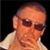 Static pixelated image of a person's head and shoulders with dark sunglasses, light skin tone, short hair, and visible ear against black background.