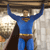 A pixelated figure in a blue suit with a yellow belt and red emblem holds a large brown beam above its head against a blurred background.