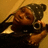 A person wearing a dark beanie with white dots holds a silver camera, positioned against a light brown background.