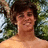 A pixelated static image shows a person with dark curly hair and light skin, wearing no shirt, against a blurred green background.