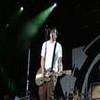 A person in a white shirt and brown pants stands on a stage holding a light-colored electric guitar under green stage lighting.