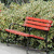 A static image shows a red bench with horizontal slats and dark legs, positioned on a grassy area with blurred green foliage in the background.