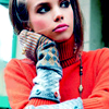Static image of a person wearing an orange turtleneck sweater and patterned arm warmer, resting their head on one hand with visible blush and makeup.