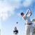 Pixelated static image of a golf icon showing a golfer mid-swing against a light blue sky background with faint cloud patterns and small dark dots resembling birds or debris.