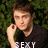 Pixelated image of a person with short brown hair wearing a black shirt, hand near face, against green foliage background.