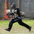 A person in dark clothing and helmet runs across grass with a paintball gun, then stands holding it horizontally.