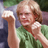 Pixelated static image of a person with light brown hair wearing a green shirt, holding an object resembling a beer mug or glass in their right hand against a blurred background.