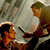 Two figures in a pixelated scene: one seated with dark hair and orange clothing, the other standing with green hair and white-and-brown attire, facing each other against a muted background.