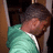 A pixelated figure in a teal shirt with white trim leans against a brown wooden structure, head tilted downward.