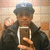 Person wearing dark jacket and blue cap holding smartphone in front of tiled wall with circular light fixture.