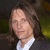 Pixelated portrait of a person with long brown hair wearing a white collared shirt and dark jacket against a blurred green and brown background.