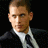 Pixelated static image of a person with short brown hair wearing a dark suit and white shirt against black background.