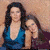 Two women in a close pose, one with long dark hair and blue top leaning toward another woman in purple top resting head on shoulder.