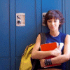 A person with short dark hair sits in front of blue lockers, holding a red book and yellow backpack, while the word REBEL appears in red digital text above.