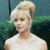 A pixelated image of a person with blonde hair styled in an updo wearing a white sleeveless top against a dark green background.