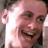 Pixelated close-up of a smiling person with light skin tone, dark hair, and visible ear, wearing a white shirt against a dark background.