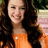 A pixelated close-up of a person with long dark hair wearing an orange sleeveless top, smiling with eyes partially closed against a blurred background.
